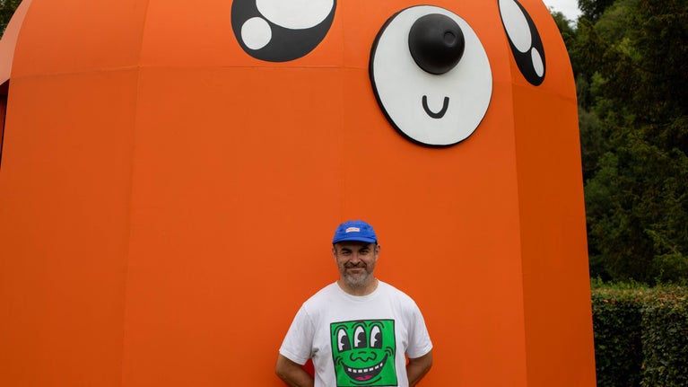An artist named Bruce Asbestos with a hat on in front of an orange sculpture of a mole